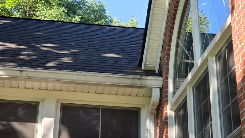 A white gutter with dark drip streaks and grime under a dark shingle roof beside an arched window.