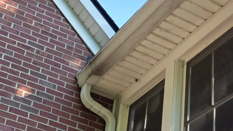 A cream gutter and downspout with dark grime along the front edge, seen from below against a brick wall.