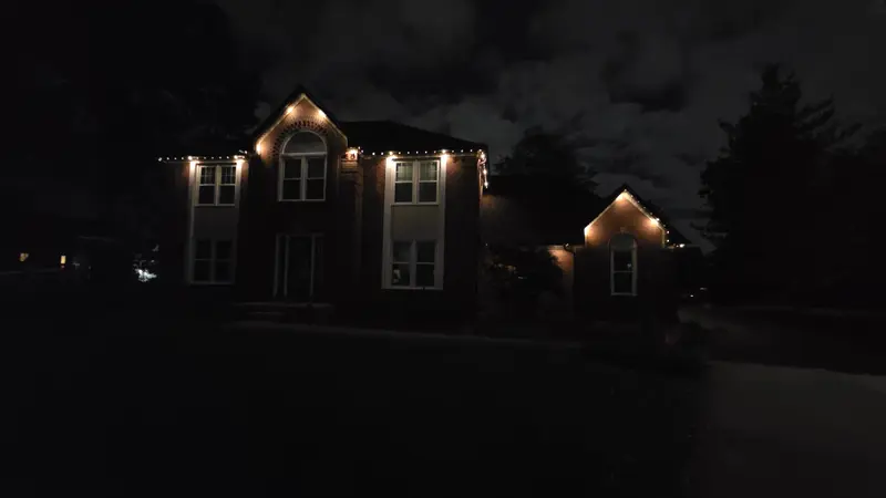A two-story brick house at night with warm white lights along the roof peaks and eaves under a cloudy sky.