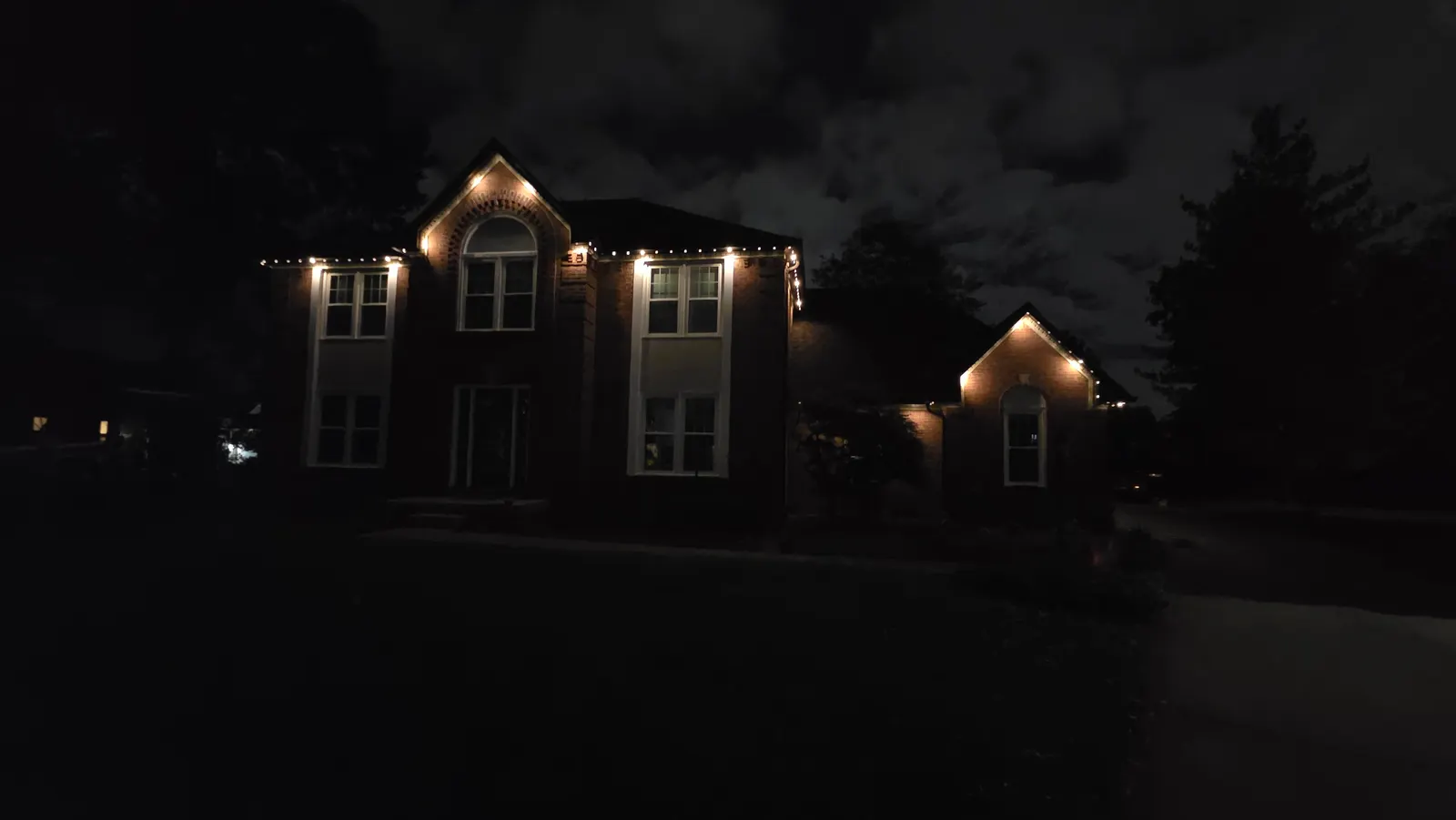 A two-story brick house at night with warm white lights along the roof peaks and eaves under a cloudy sky.