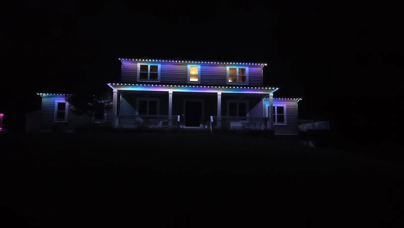 A two-story house with a front porch at night, its rooflines and porch outlined in blue and teal permanent LED lights.