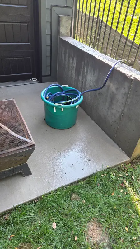 The same concrete patio corner beside the dark garage door, now clean light gray concrete, still wet from washing.