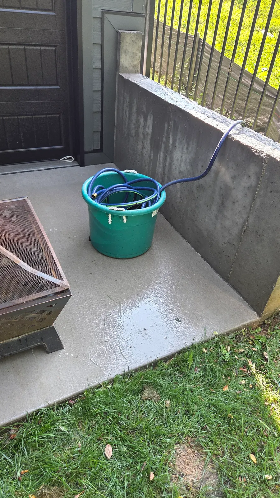 The same concrete patio corner beside the dark garage door, now clean light gray concrete, still wet from washing.