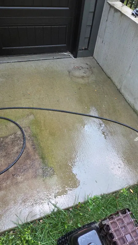 Concrete patio corner beside a dark garage door, stained green and brown with algae, with a pressure hose lying across it.