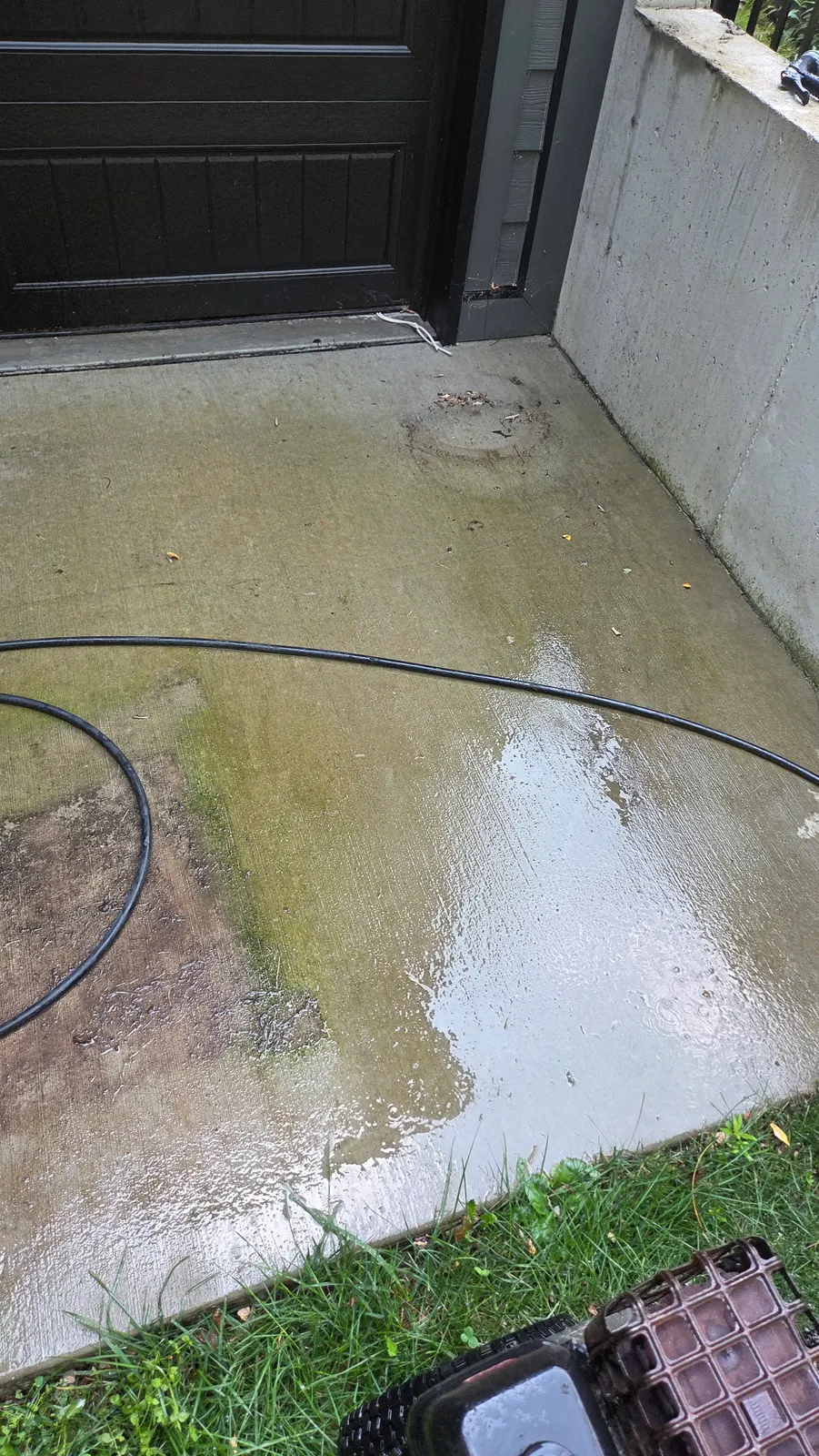 Concrete patio corner beside a dark garage door, stained green and brown with algae, with a pressure hose lying across it.