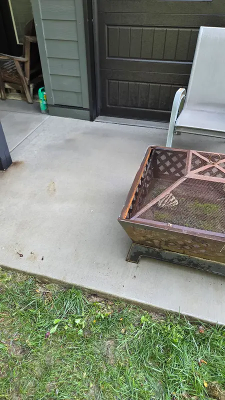 The same concrete slab in front of the dark garage door, clean and even in color, with a fire pit and chair set back on it.