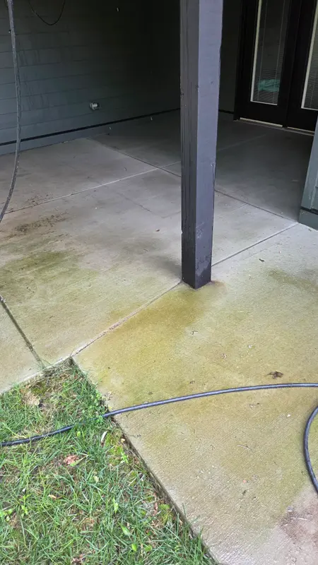 Concrete patio around a dark support post with green algae staining along the edge by the lawn.