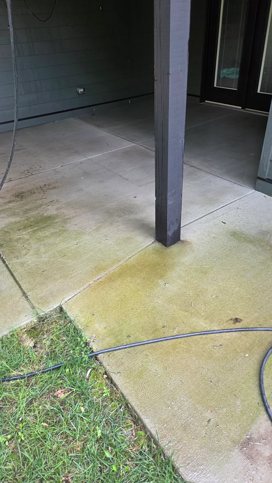Concrete patio around a dark support post with green algae staining along the edge by the lawn.