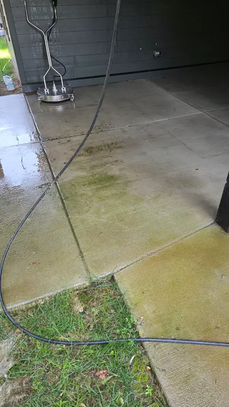 A surface cleaner and pressure hose on a concrete patio that is half cleaned, with green algae still on the unwashed section.