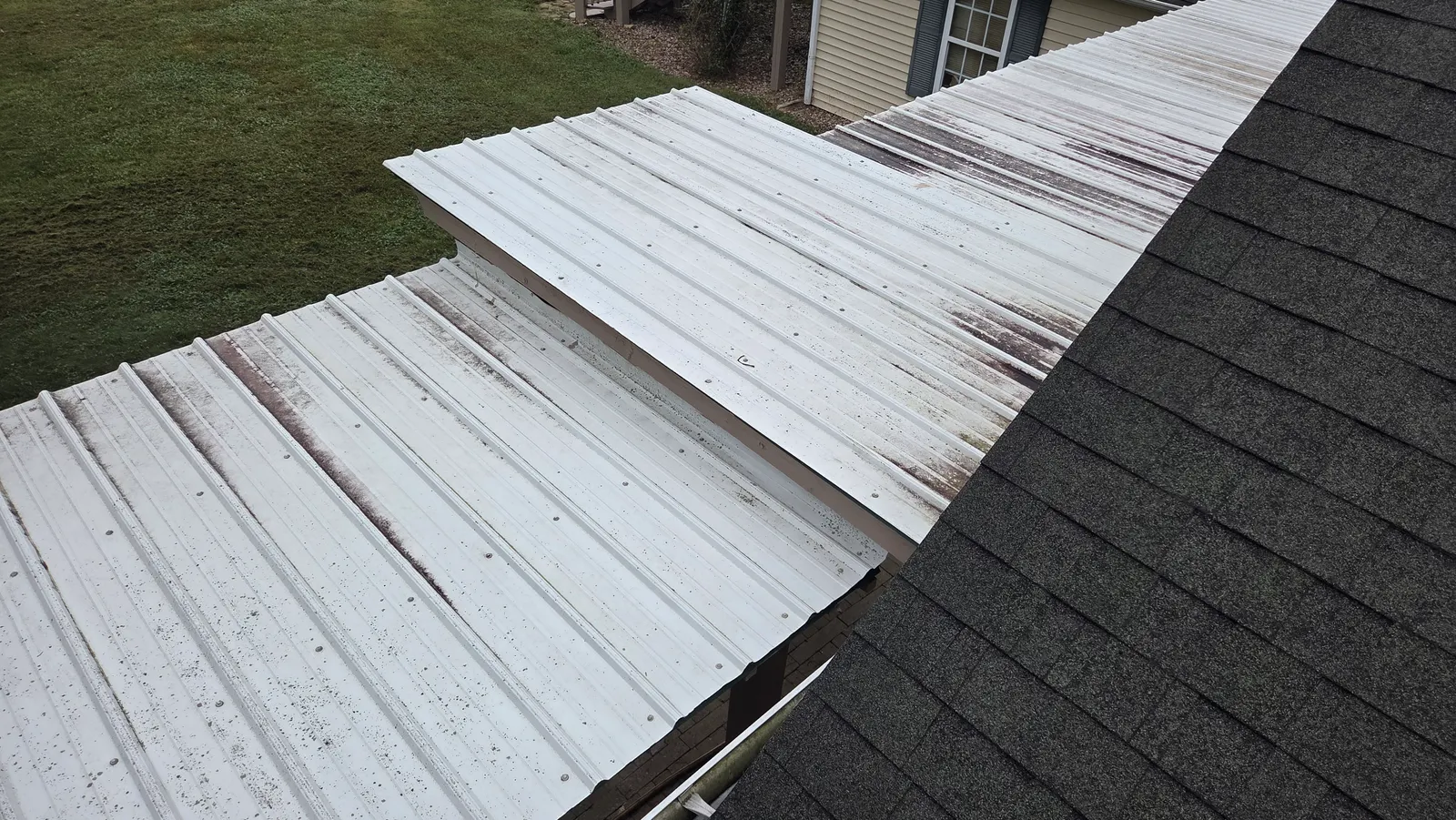 A white metal porch roof with dark rust-colored streaks running down the panels, next to dark shingles.