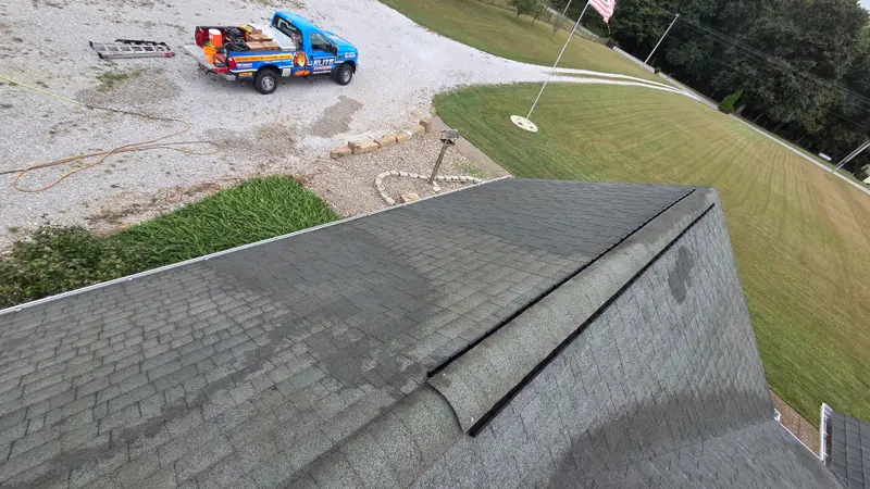 The same roof ridge partway through washing with lighter cleaned shingles, and the blue Elite Exteriors truck parked in the gravel drive below.