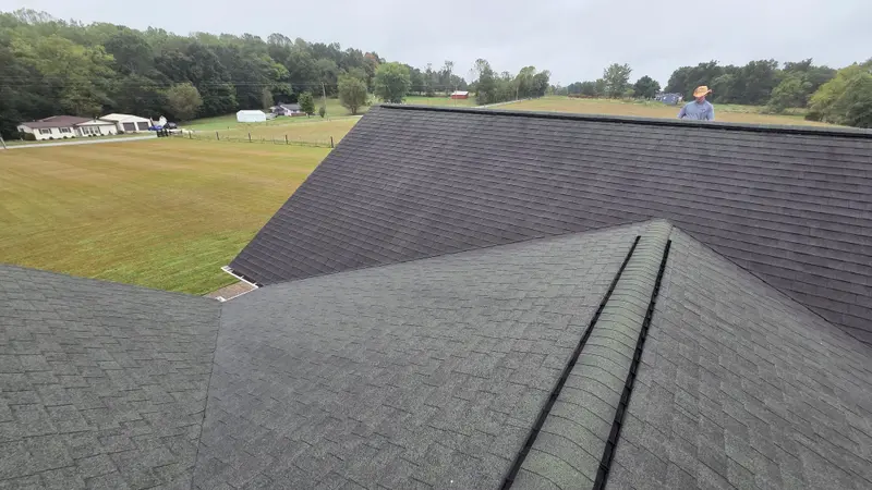 A wide view of a dark, streaked shingle roof with open fields and trees behind it.