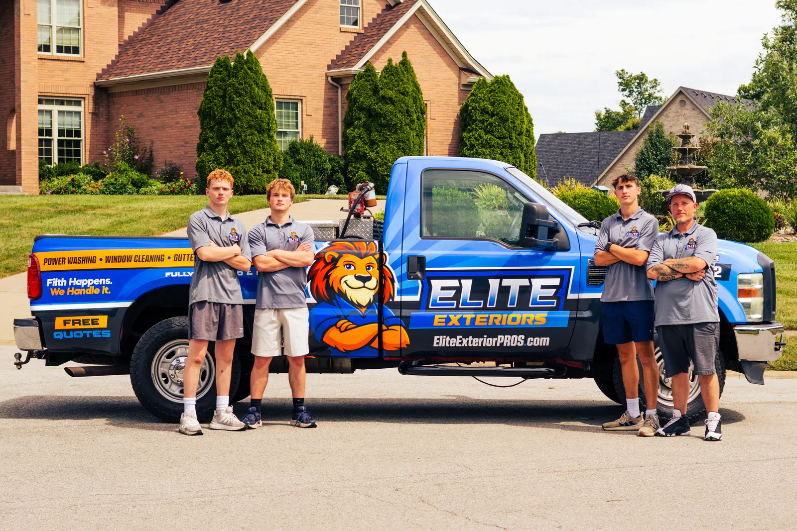Four crew members in gray Elite Exteriors polos stand with arms crossed beside the blue wrapped Elite Exteriors pickup truck on a residential street.