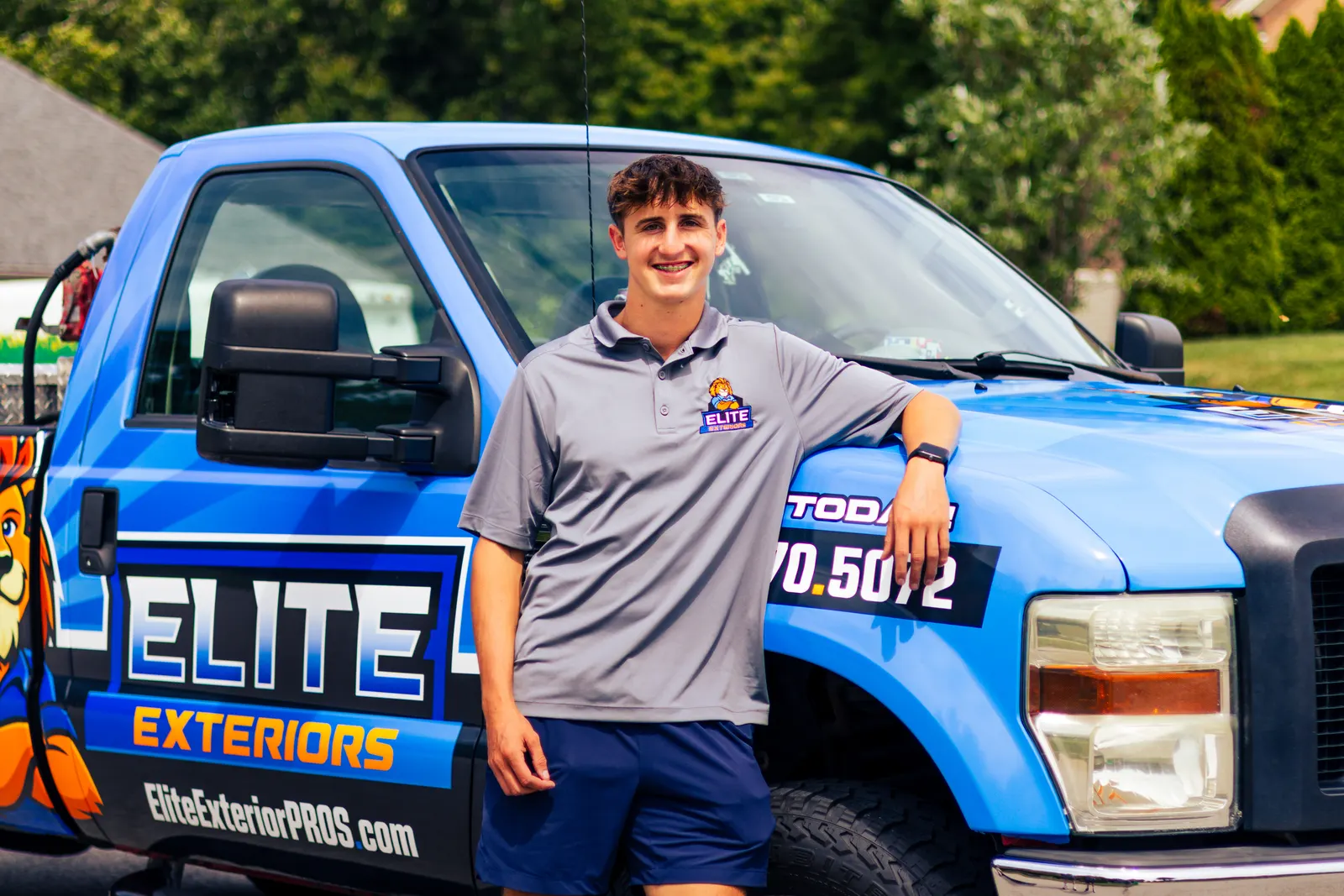 A smiling dark-haired young man in a gray Elite Exteriors polo leans on the hood of the blue wrapped Elite Exteriors pickup truck.