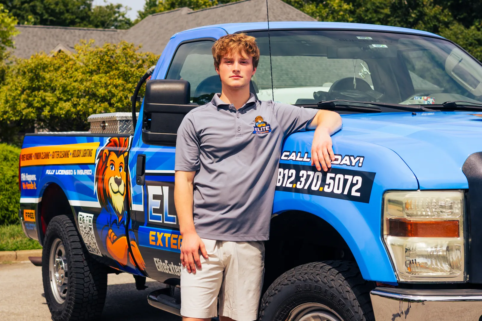 A young man with wavy brown hair in a gray Elite Exteriors polo leans on the hood of the blue wrapped Elite Exteriors pickup truck.