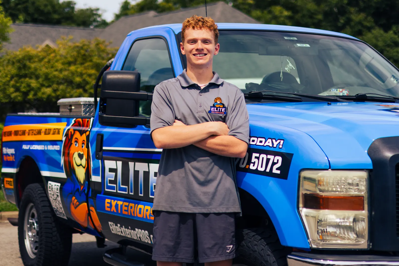 A smiling red-haired young man in a gray Elite Exteriors polo stands with arms crossed in front of the blue wrapped Elite Exteriors pickup truck.
