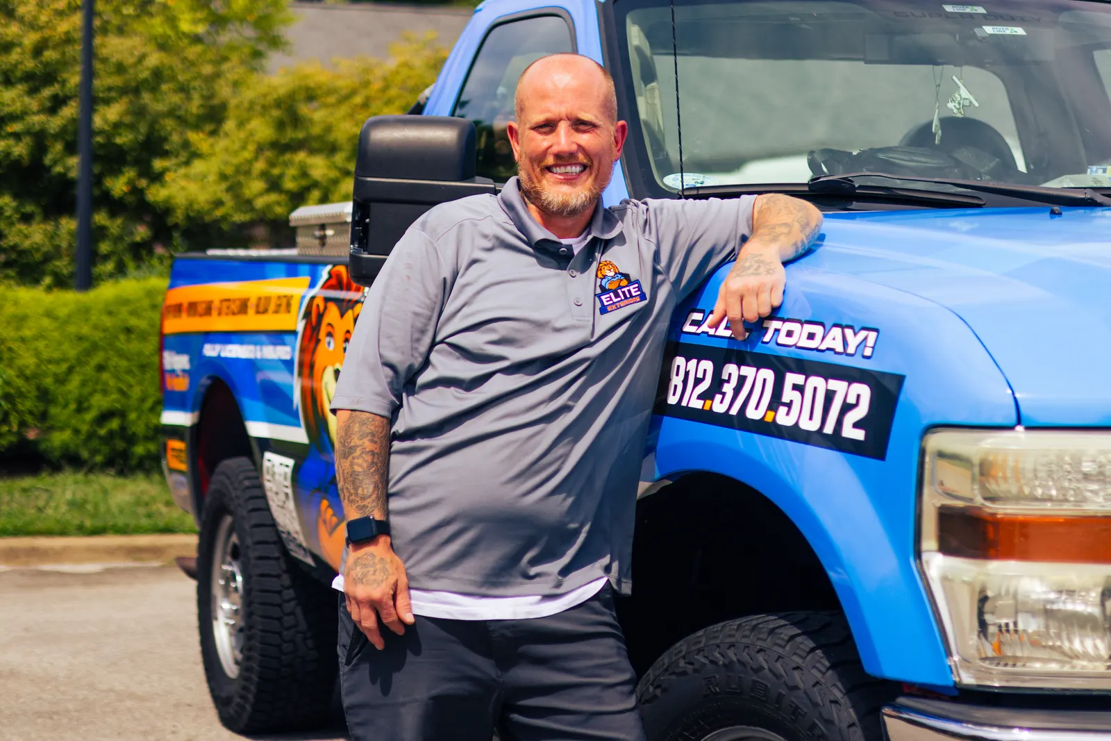 A smiling bald, bearded man with tattooed forearms in a gray Elite Exteriors polo leans on the hood of the blue wrapped Elite Exteriors pickup truck.