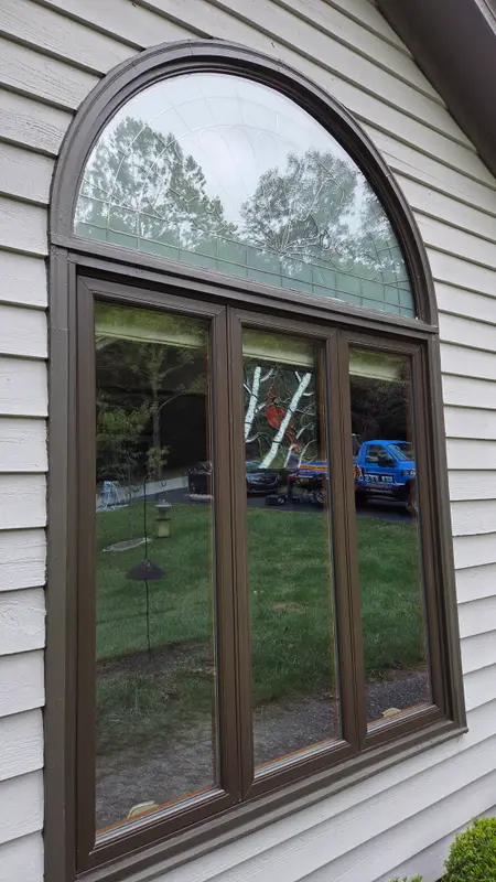 A tall brown-framed window with an arched top on a gray sided house, the glass reflecting the yard and the blue Elite Exteriors truck.