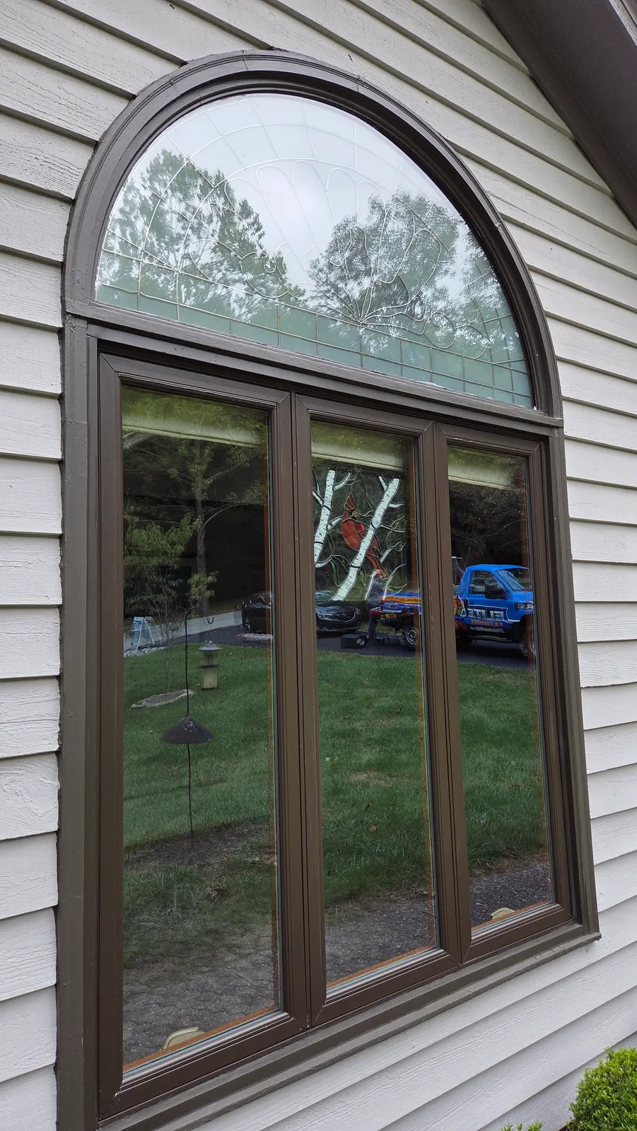 A tall brown-framed window with an arched top on a gray sided house, the glass reflecting the yard and the blue Elite Exteriors truck.