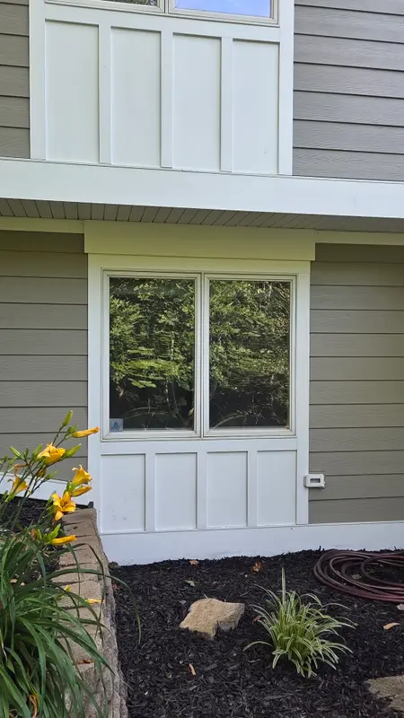 A white-trimmed double window with clear glass on a taupe sided house above a mulched flower bed with daylilies.