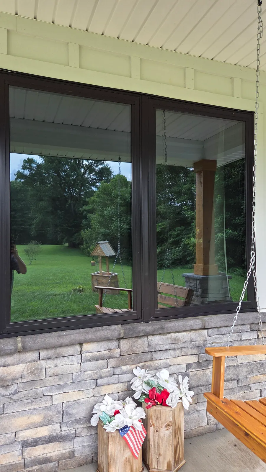 Clean dark-framed double window on a covered front porch with stone veneer below, reflecting the yard.