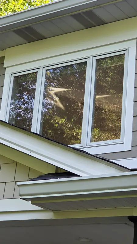 A white-trimmed triple window on the upper story of a taupe house, the clean glass reflecting trees in sunlight.